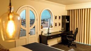 A hotel room with a bed, a desk with a computer, a chair, and two large arched windows offering a view of the city. - Corona Hotel & Spa (Ensenada)