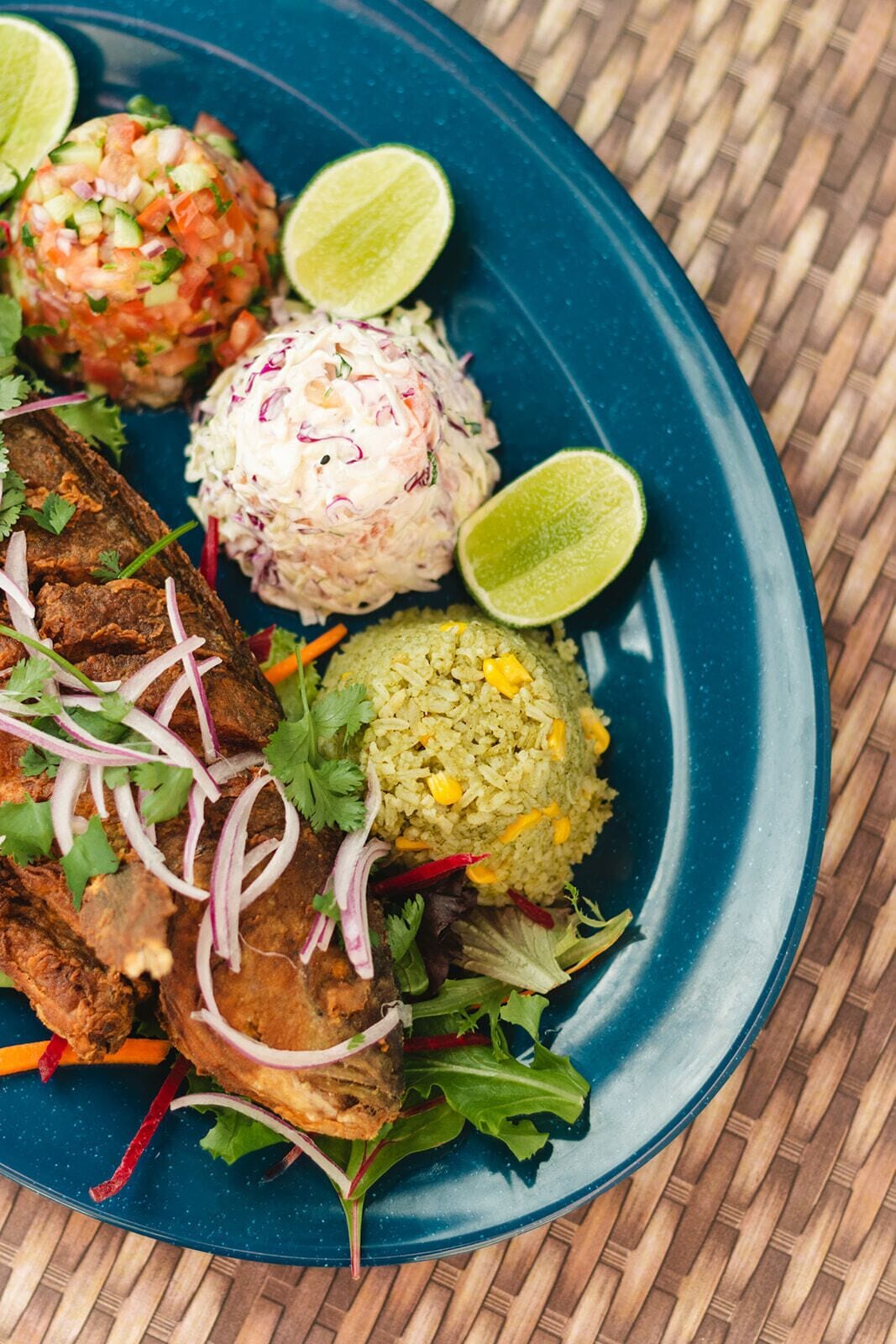 A plate with a fried fish sandwich, coleslaw, and rice balls, garnished with cilantro and lime wedges.