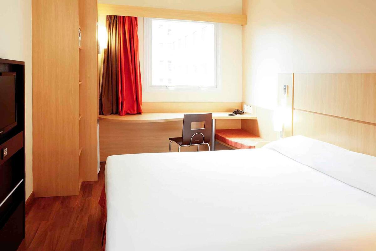 A hotel room with a bed, a desk, a chair, a TV, and a window with curtains.