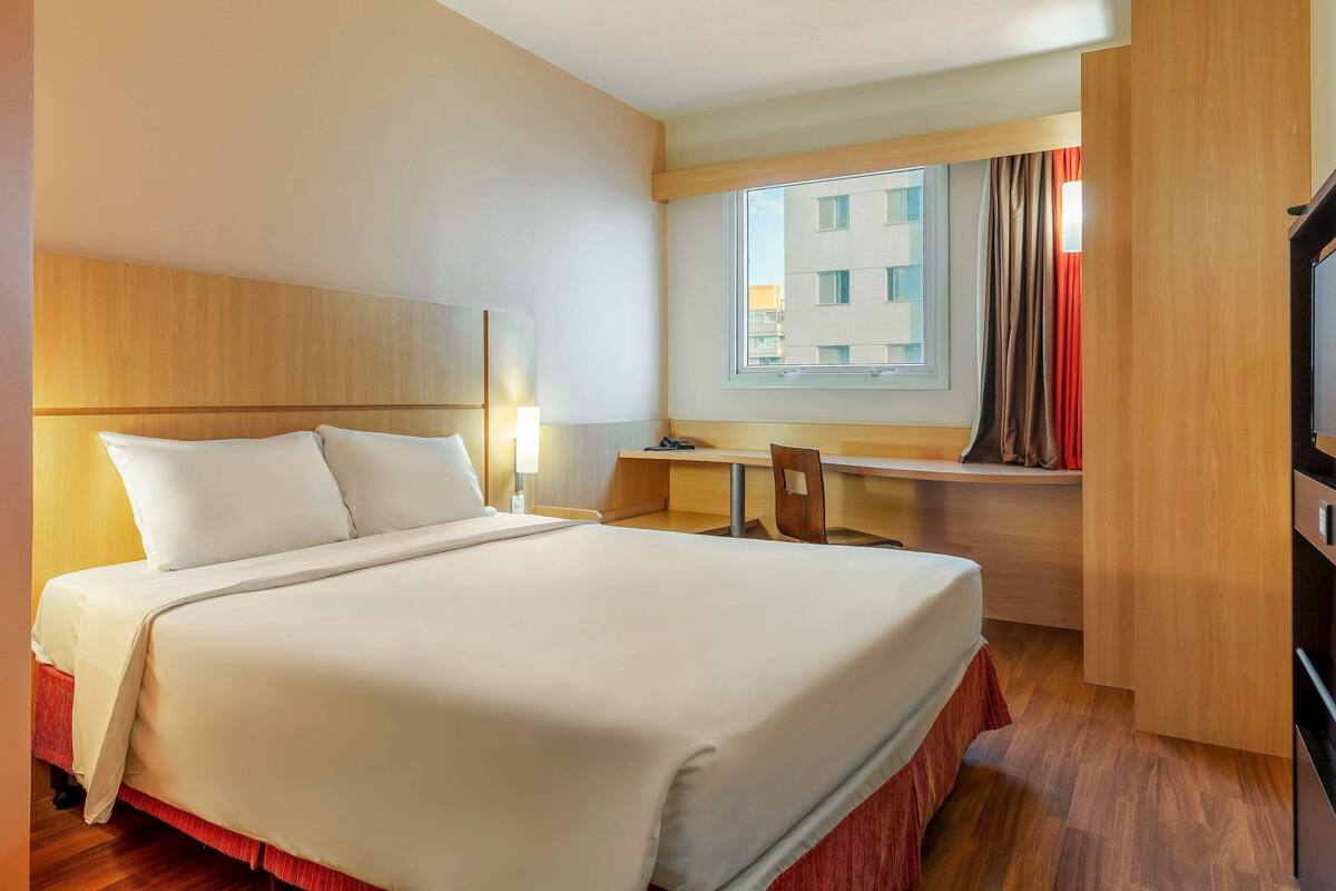 A hotel room with a bed, a desk, a chair, a TV, and a window with curtains.