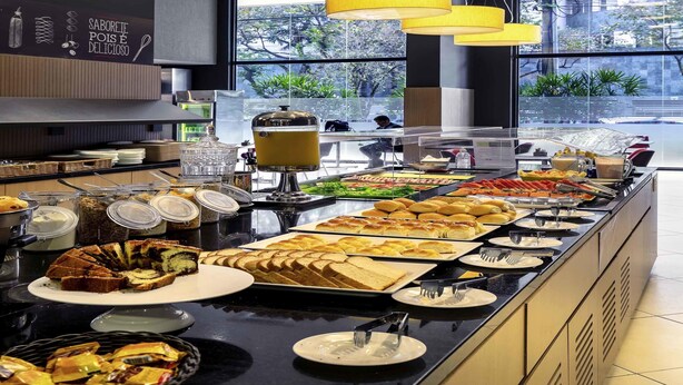 A buffet area with various food items and a coffee station, with large windows and natural light. - ibis BH Afonso Pena Savassi (Belo Horizonte)