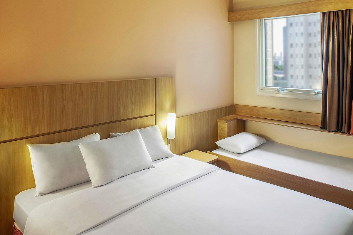 A hotel room with a large bed, a smaller bed, a nightstand, a lamp, and a window with curtains.