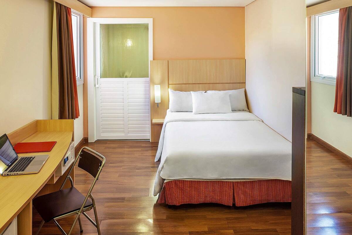 A hotel room with a bed, a desk with a laptop, a chair, and a door leading to a balcony.