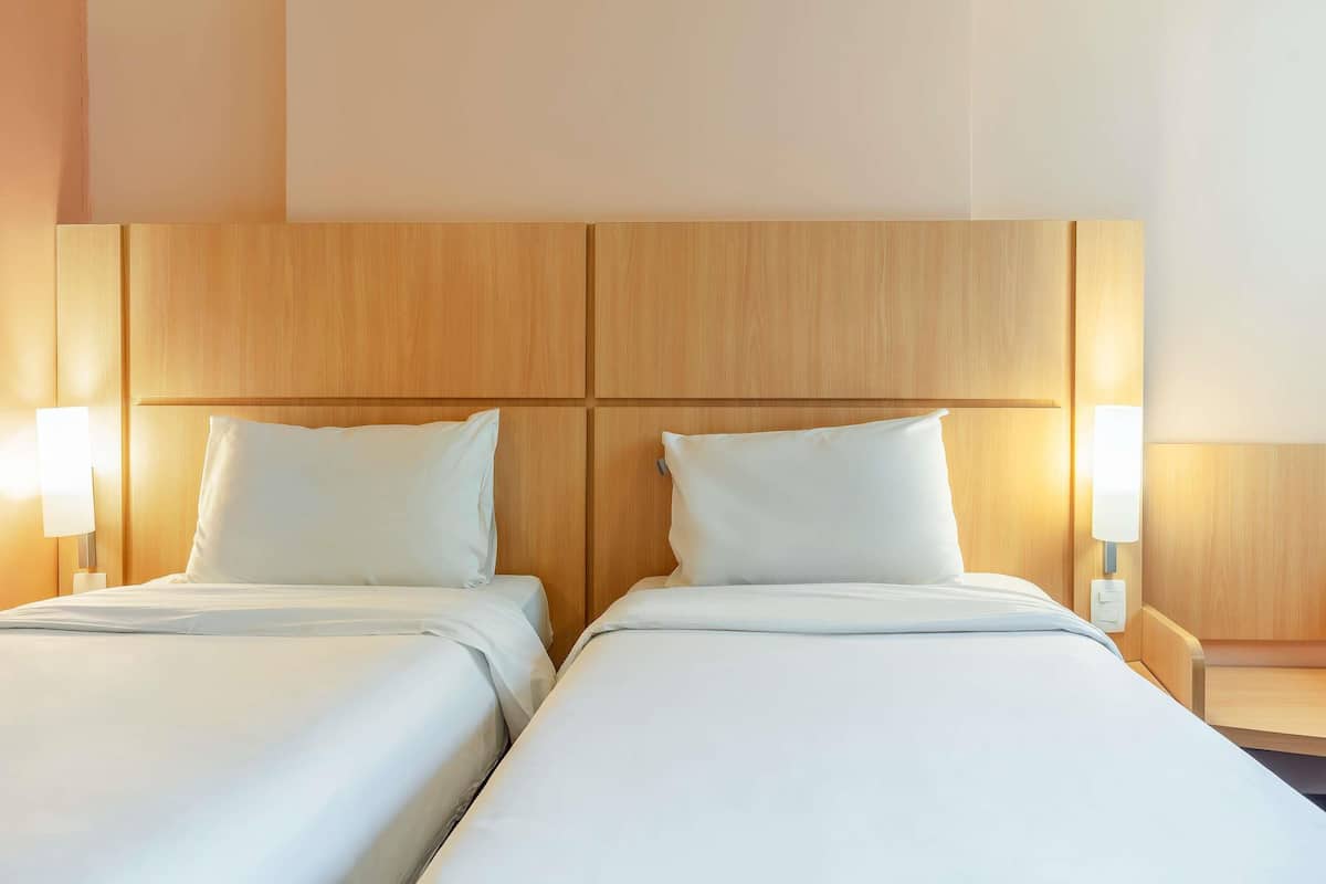 A hotel room with two beds, wooden headboard, bedside lamps, and a neutral color scheme.