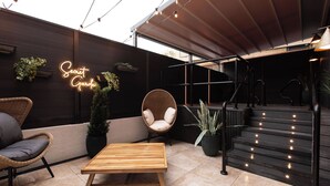 Uma área de estar moderna ao ar livre, com mesa e cadeiras de madeira, um letreiro neon e uma escadaria iluminada.