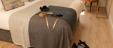 A neatly made bed with a grey blanket, a pair of black shoes, and a camera on the floor.