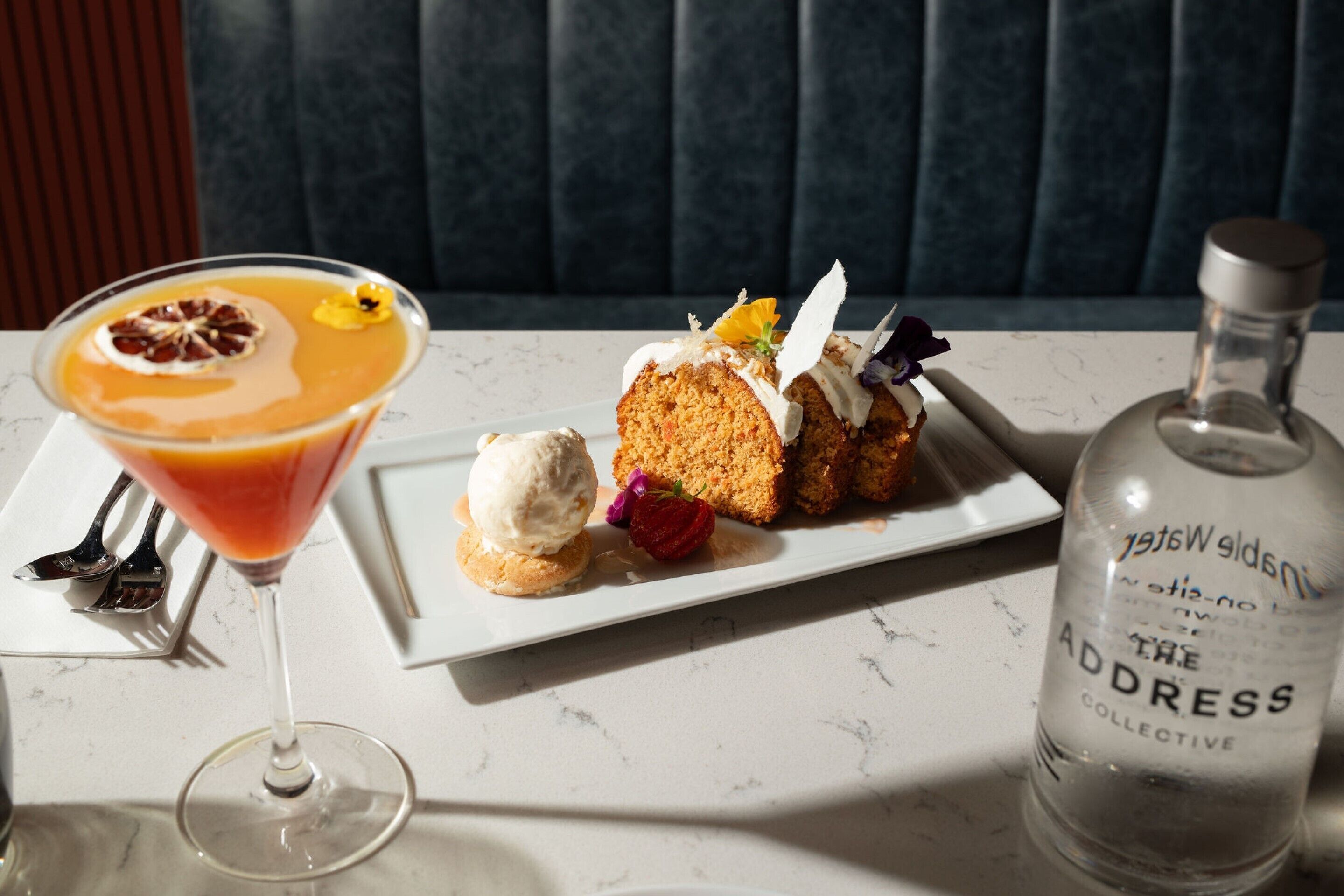 Un plato de postre con una porción de pastel, una bola de helado y una copa de cóctel con adorno. También se ve una botella de The Address Collective.