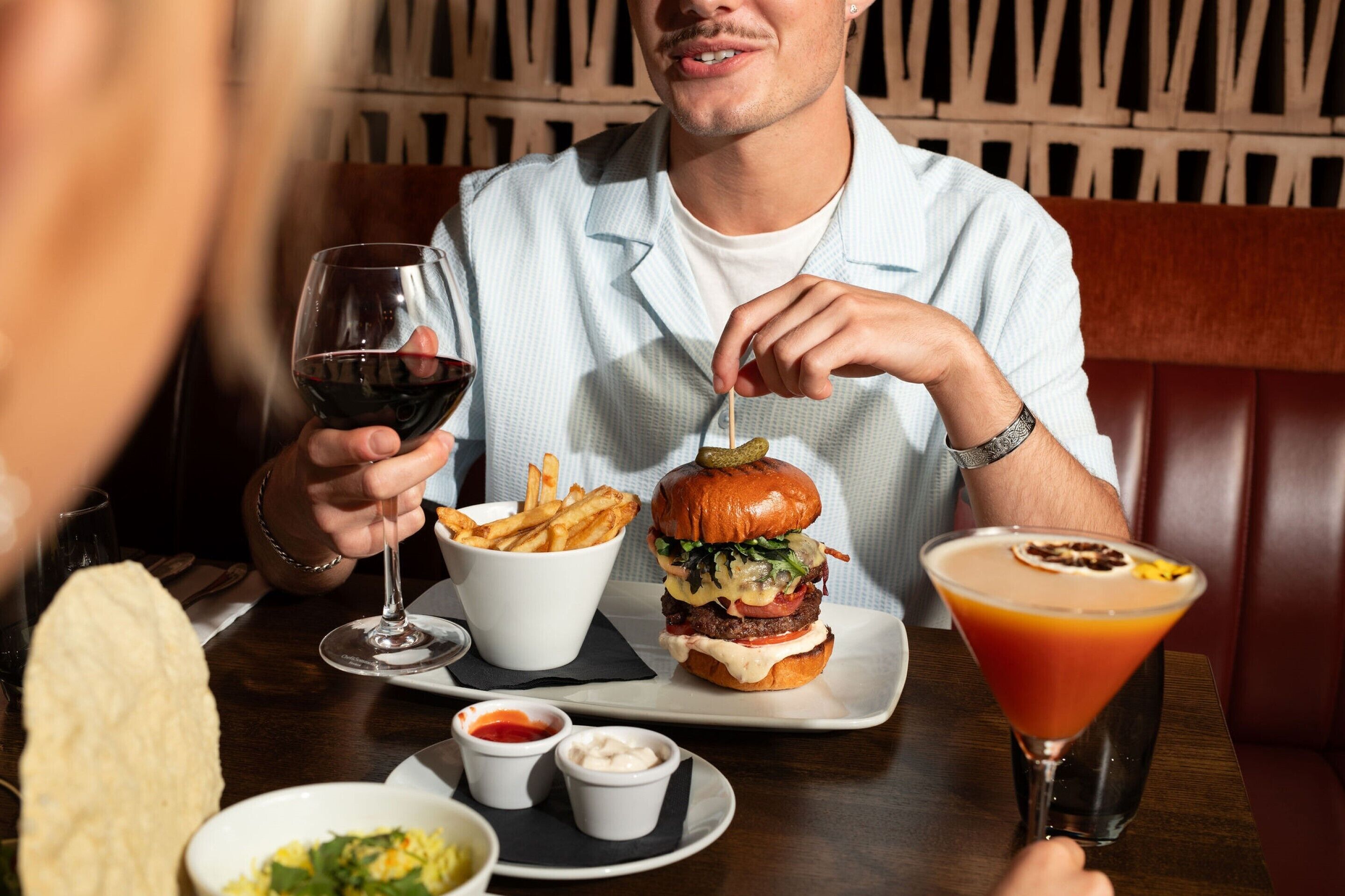 Una persona sentada a una mesa con una hamburguesa, papas fritas, un martini y recipientes pequeños de salsas.