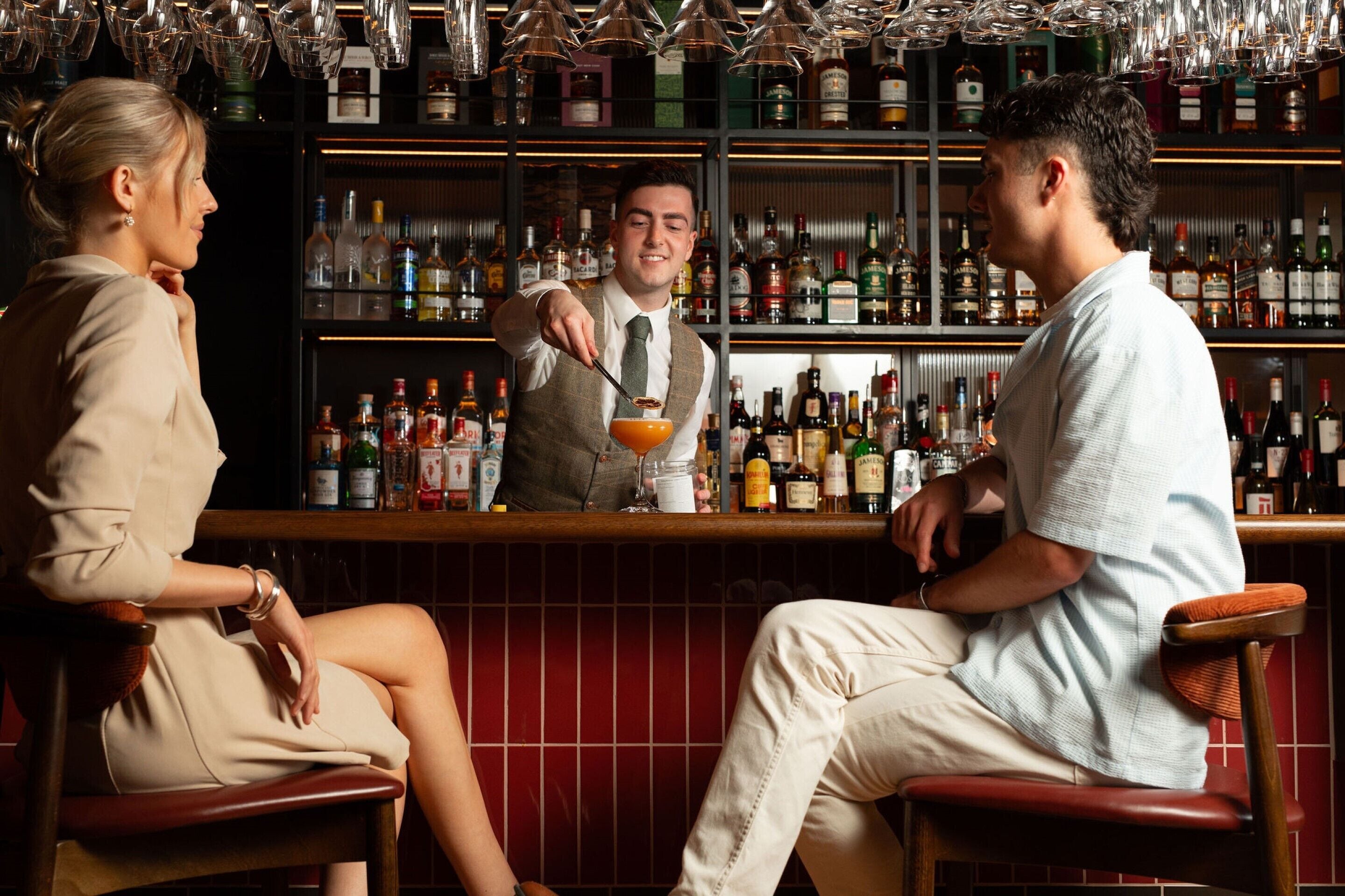 A bar area with a bartender preparing a drink, shelves stocked with various liquor bottles, and two seated customers.