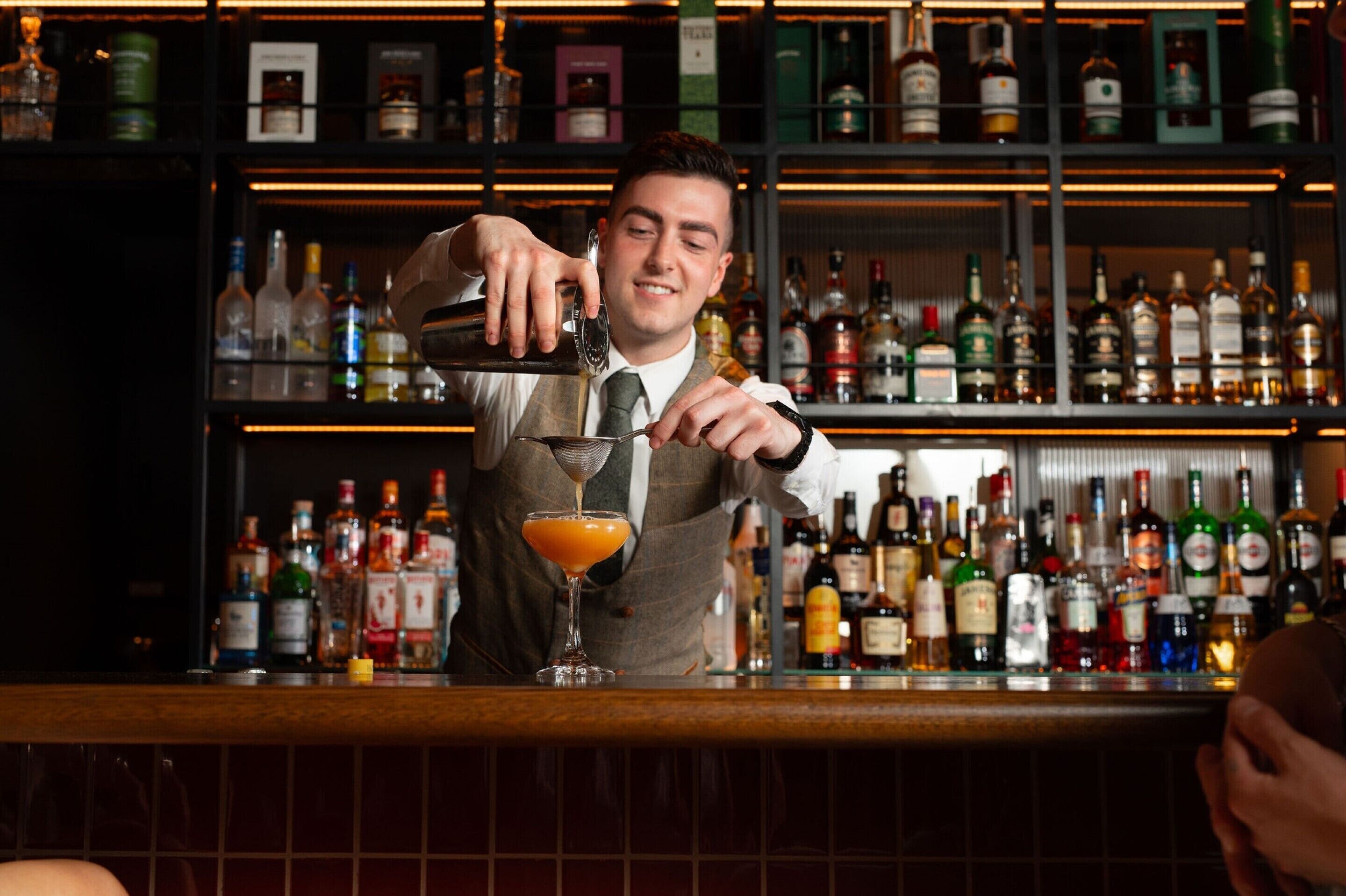 A bartender pouring a drink into a martini glass at a bar with shelves of liquor bottles in the background.
