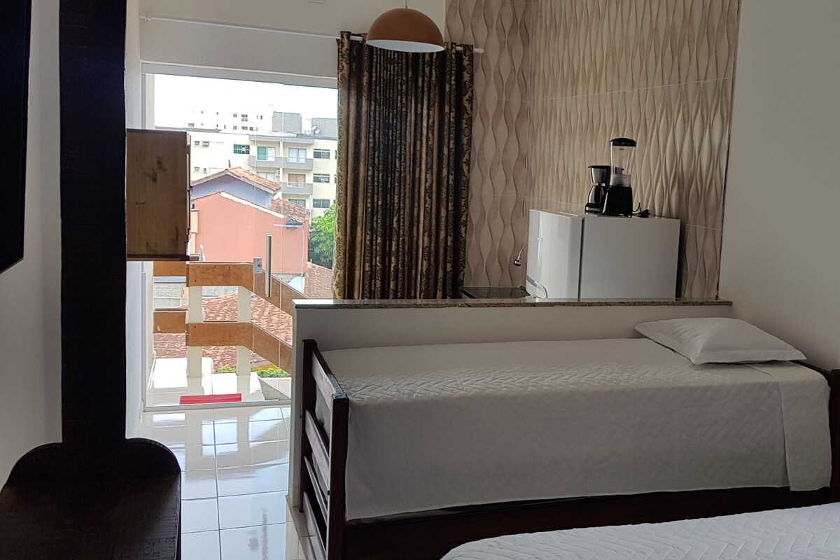 Habitación de hotel con una cama, un escritorio pequeño con cafetera, una ventana con cortinas y vistas a edificios.