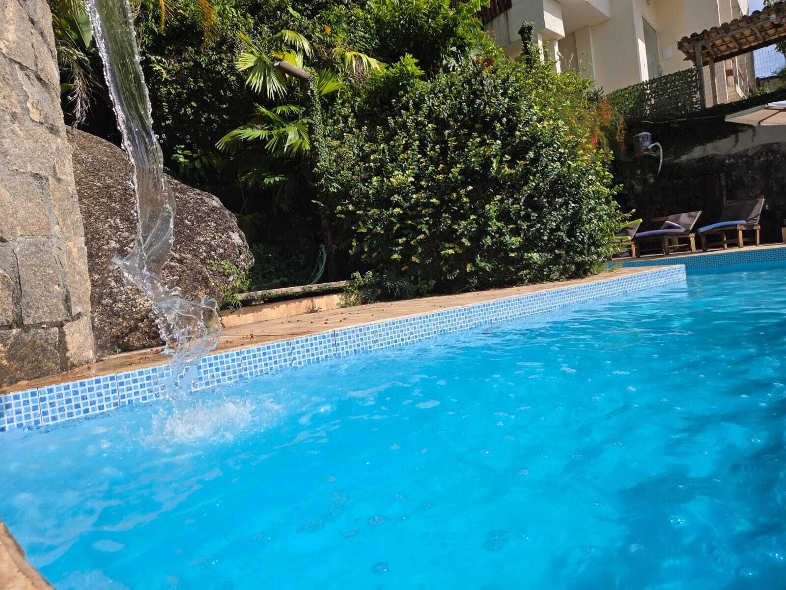 A swimming pool with a waterfall feature, surrounded by lush greenery and a tiled edge.