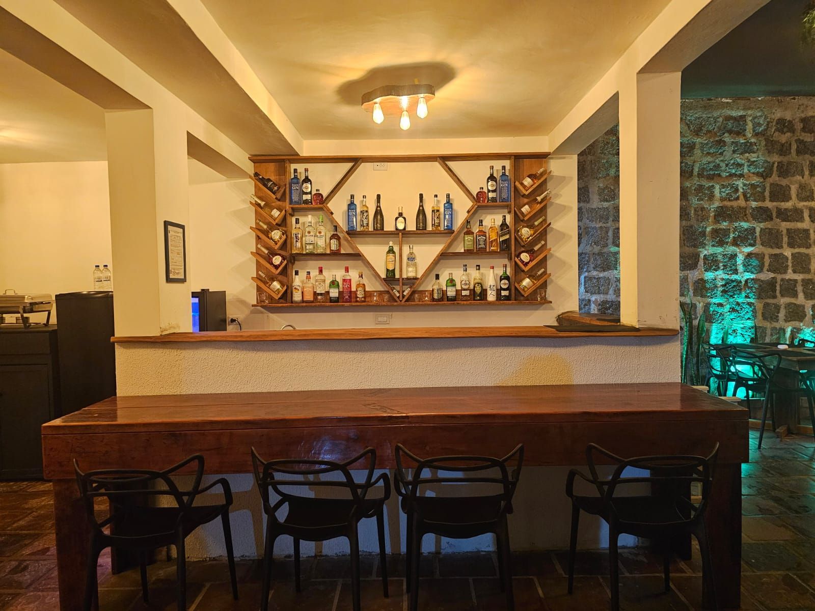 A bar area with a wooden counter and multiple shelves of liquor bottles. There are four chairs in front of the bar.