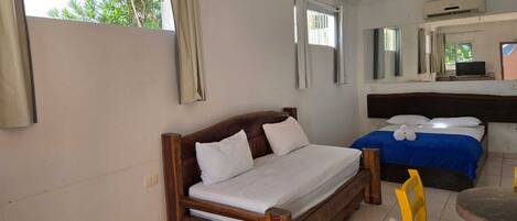 A hotel room with a wooden bed, a smaller bed with a blue blanket, a round table with a yellow chair, and a large window with curtains.
