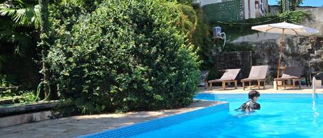 Une piscine avec une personne dans l’eau, entourée d’une végétation luxuriante et d’un bâtiment doté d’un toit en tuiles.