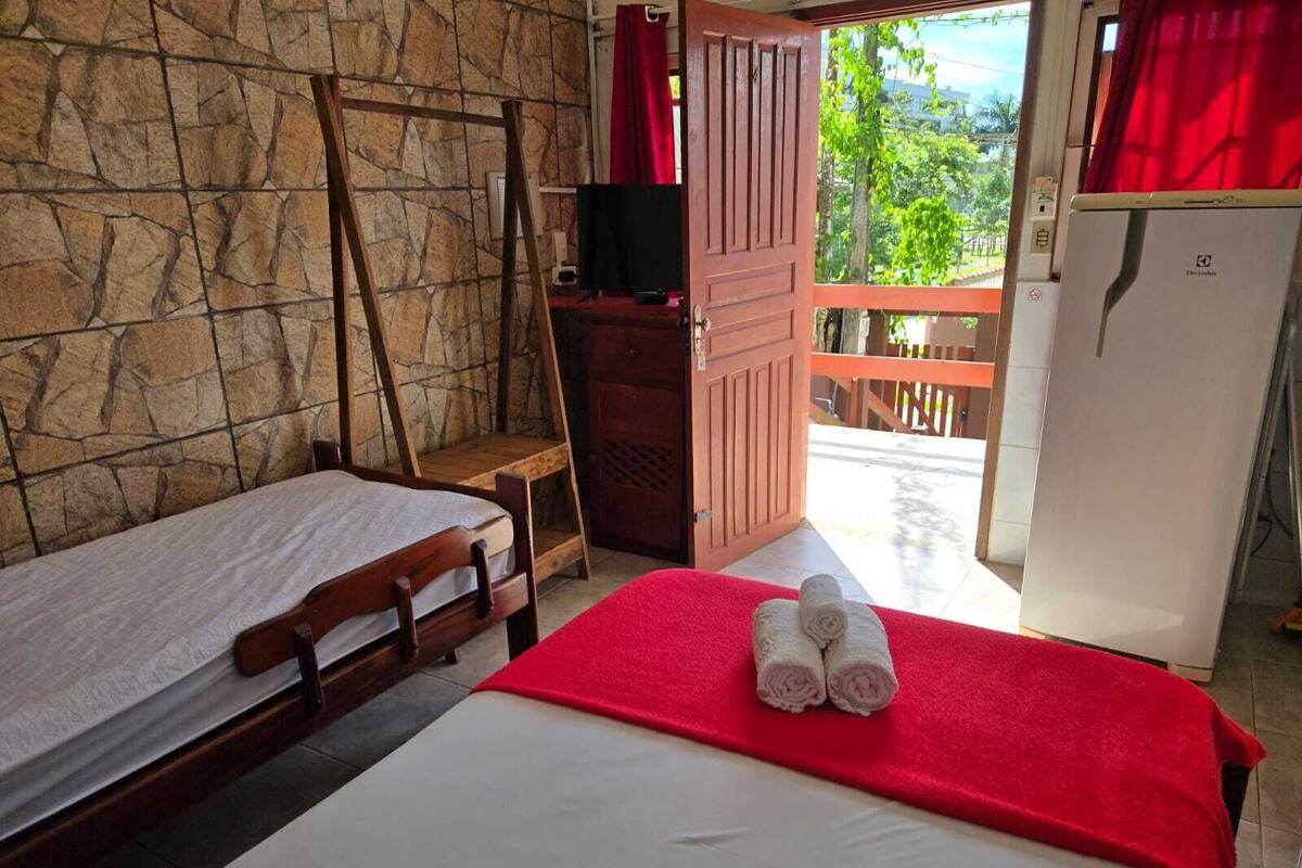 A room with a bed, a wooden bench, a stone wall, a red door, and a view of the outside.