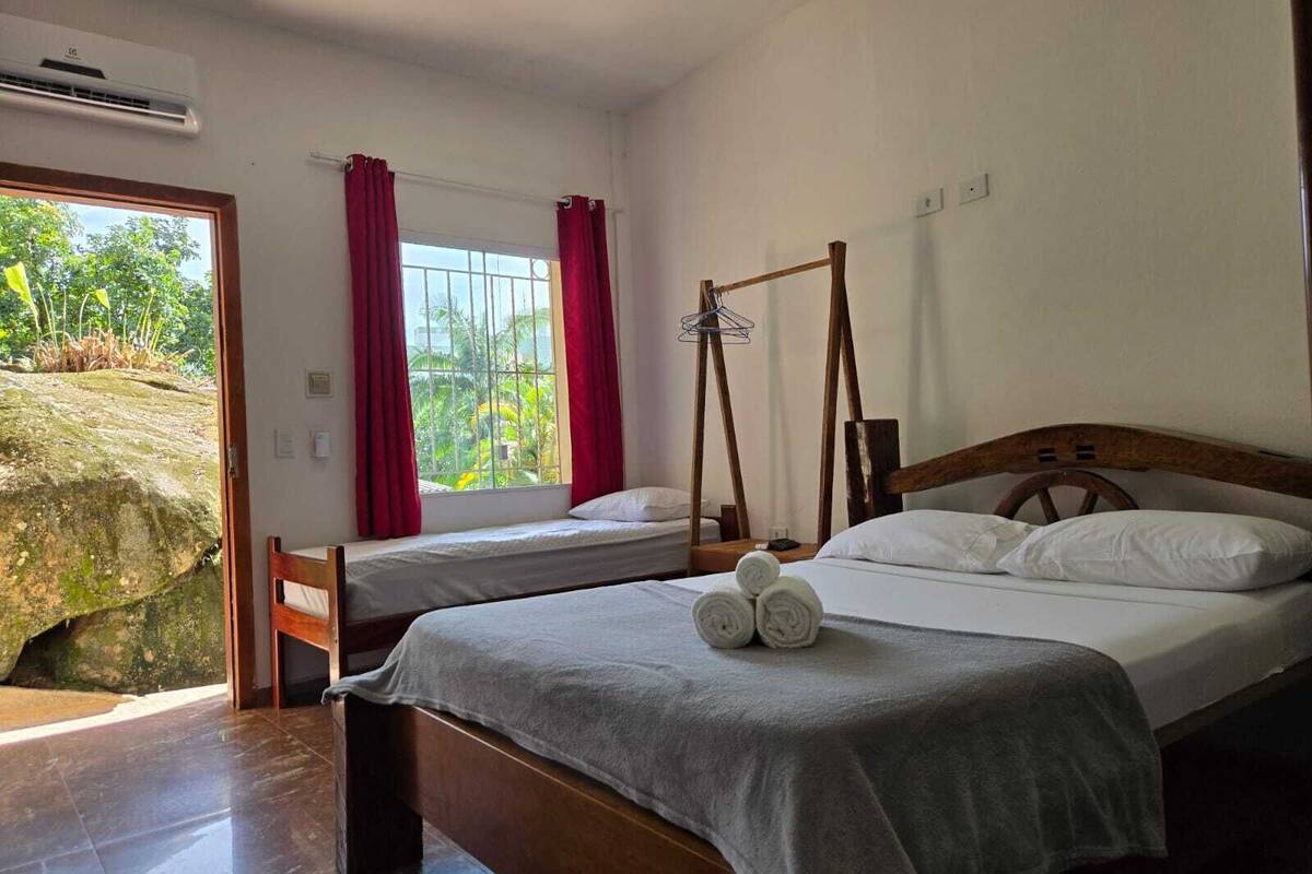 A bedroom with two beds, a wooden headboard, a window with red curtains, and a view of greenery outside.