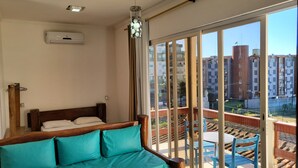 A bedroom with a wooden bed, teal pillows, and a view of a balcony with blue chairs and a railing. - Apartamentos Casa Da Pedra (Ubatuba)