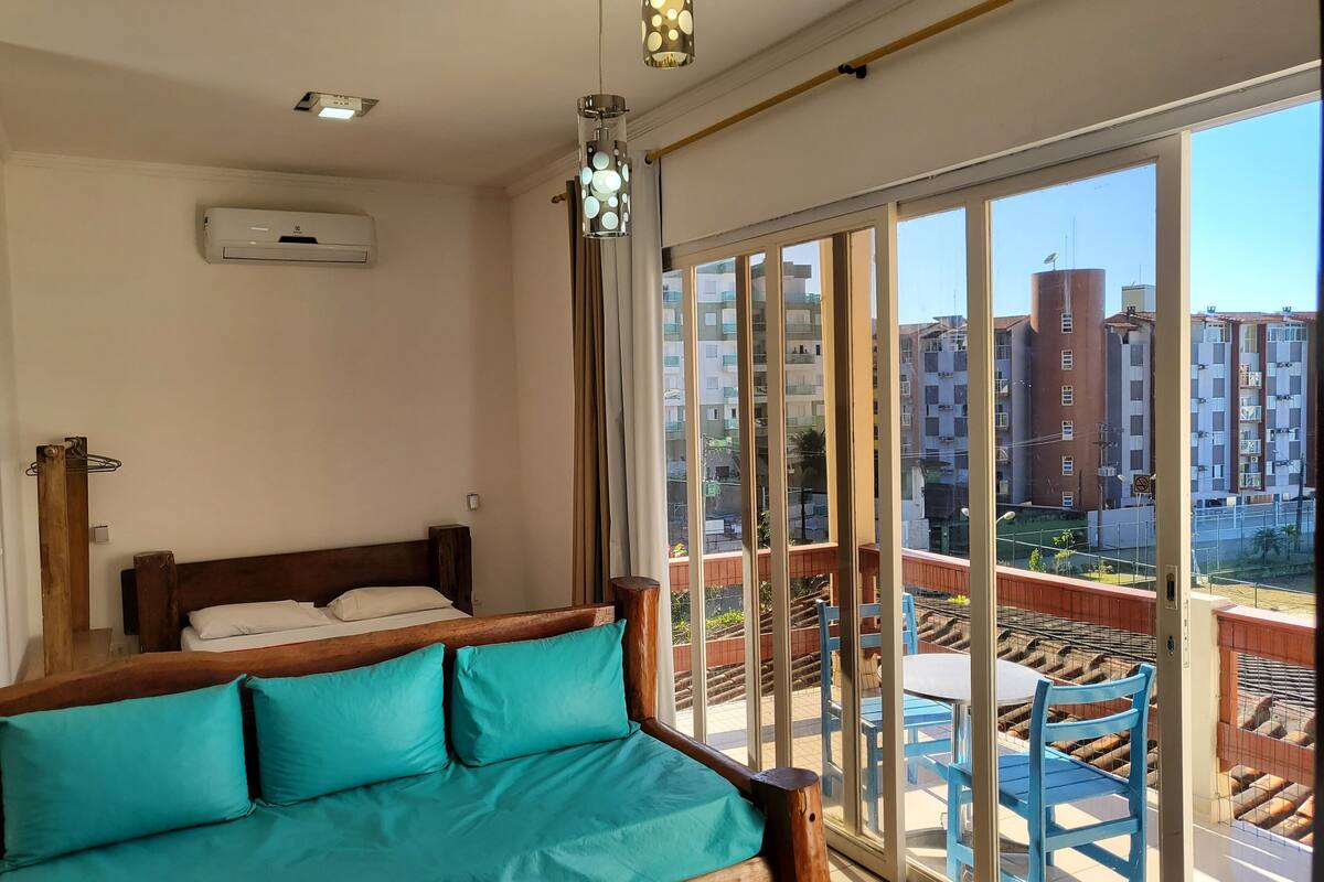 A bedroom with a wooden bed, teal pillows, and a view of a balcony with blue chairs and a railing.
