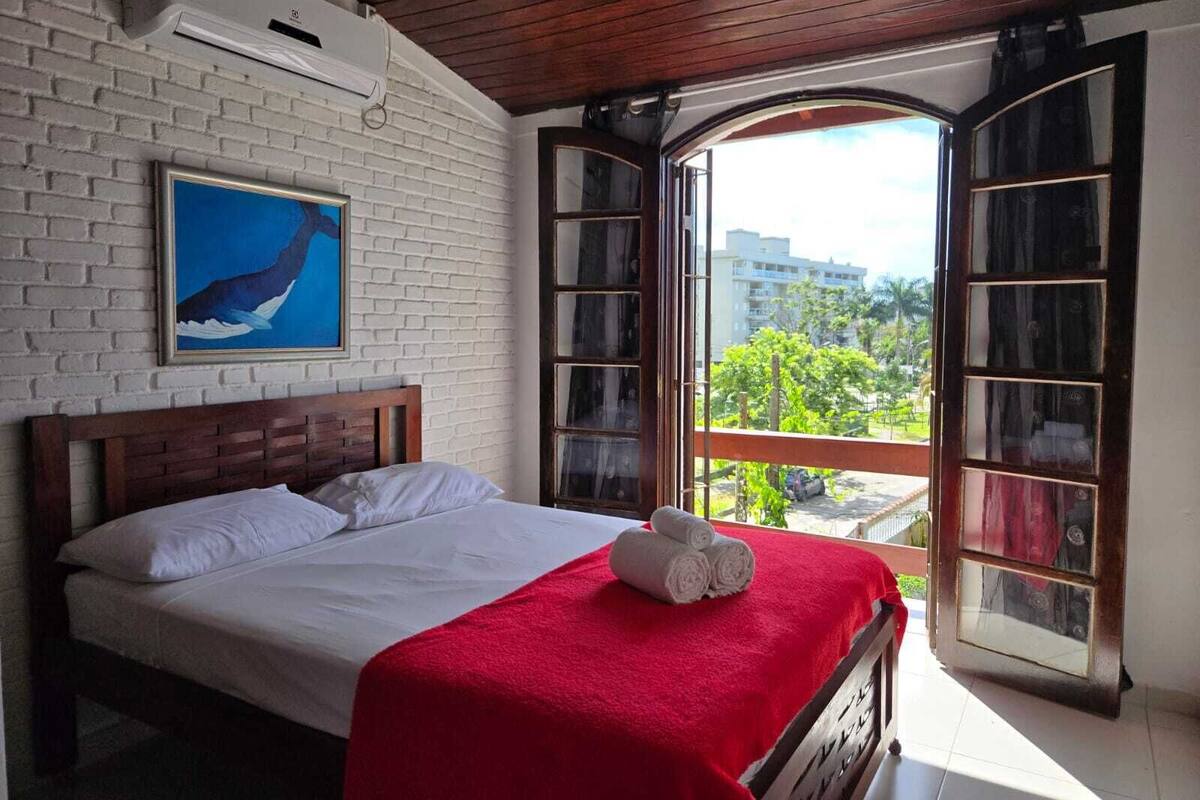 A bedroom with a bed, a red blanket, a painting of a whale, and a view of buildings and trees through open windows.