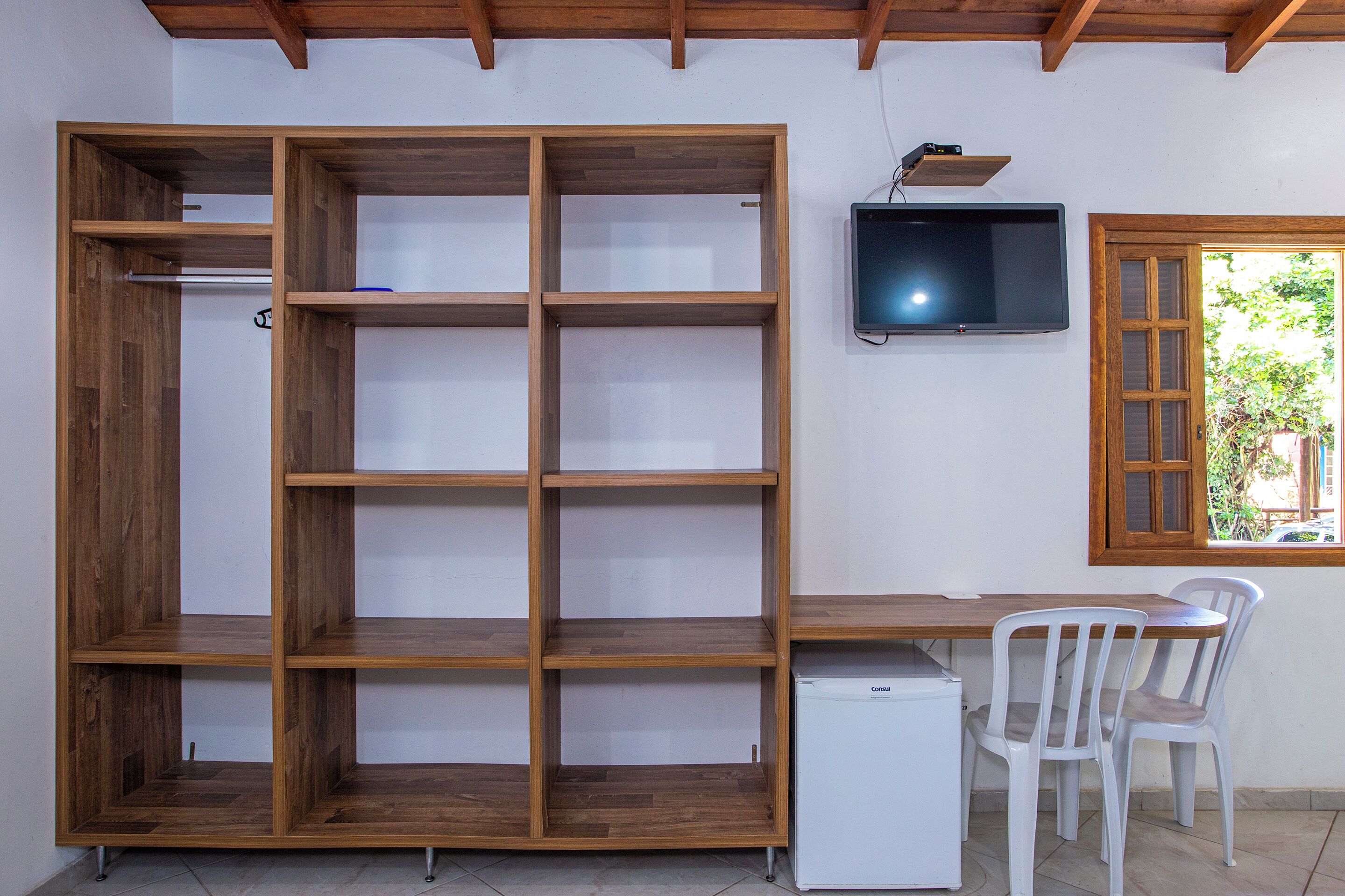 A room with a wooden shelving unit, a small white refrigerator, two white chairs, and a television mounted on the wall.