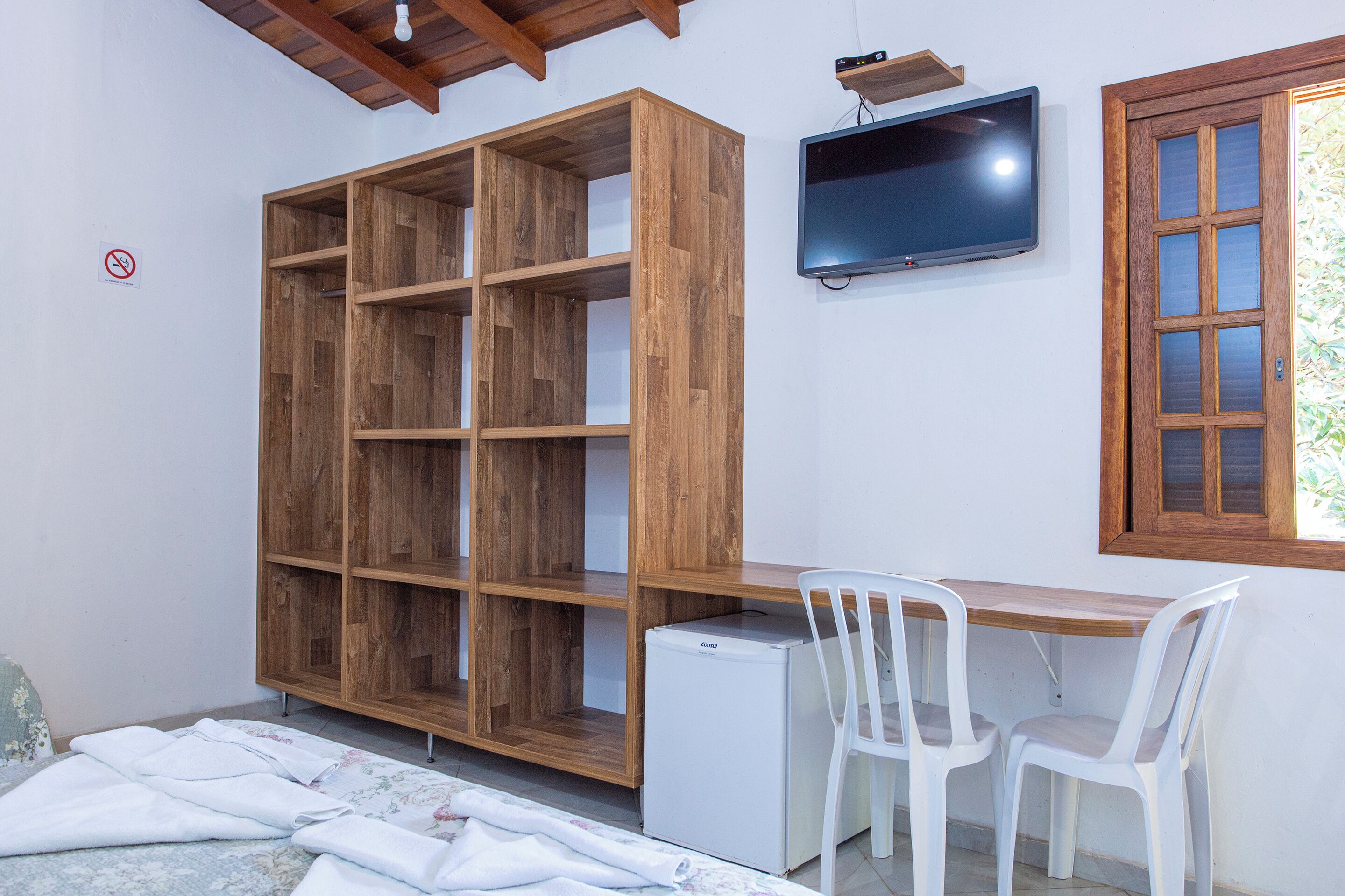 Una habitación con un armario de madera, un refrigerador pequeño, un escritorio con dos sillas blancas y un televisor montado en la pared.