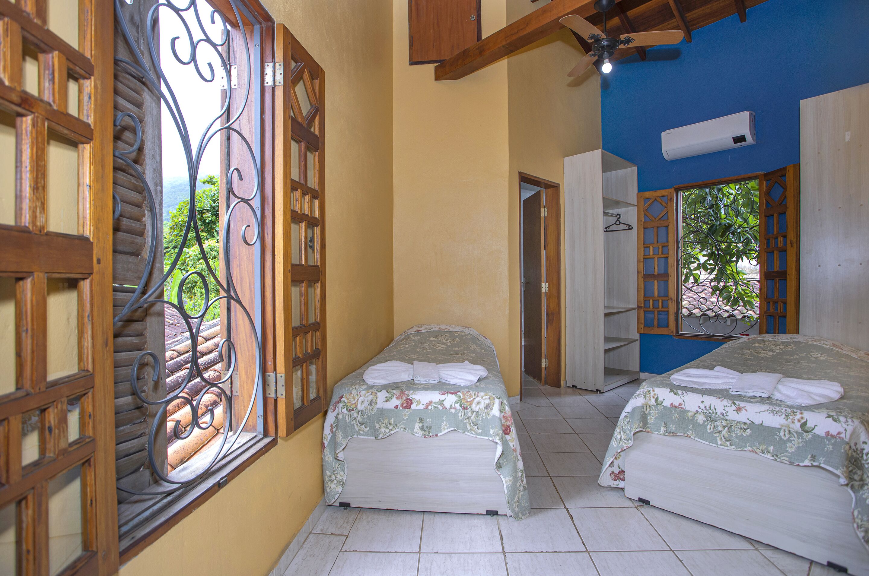 A room with two beds, a wooden door with a decorative metal design, and a view of greenery through the door.