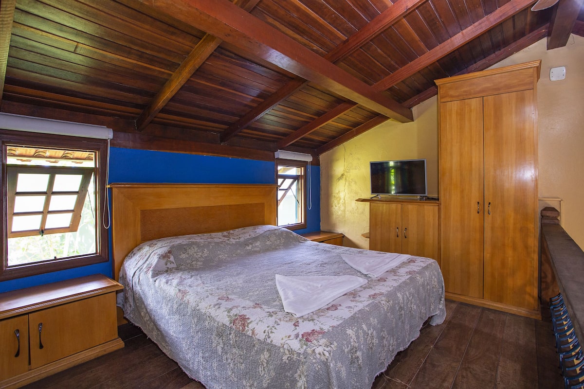 A bedroom with a wooden ceiling, a bed with a floral bedspread, a wooden wardrobe, and a television on a shelf.