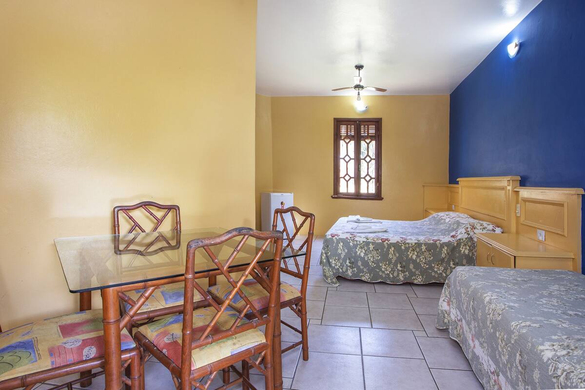 A small, compact room with a dining table, two chairs, a bed with floral bedding, a wooden nightstand, and a ceiling fan.