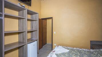 Una habitación con cama, un refrigerador pequeño, repisas de madera y un televisor montado en la pared.