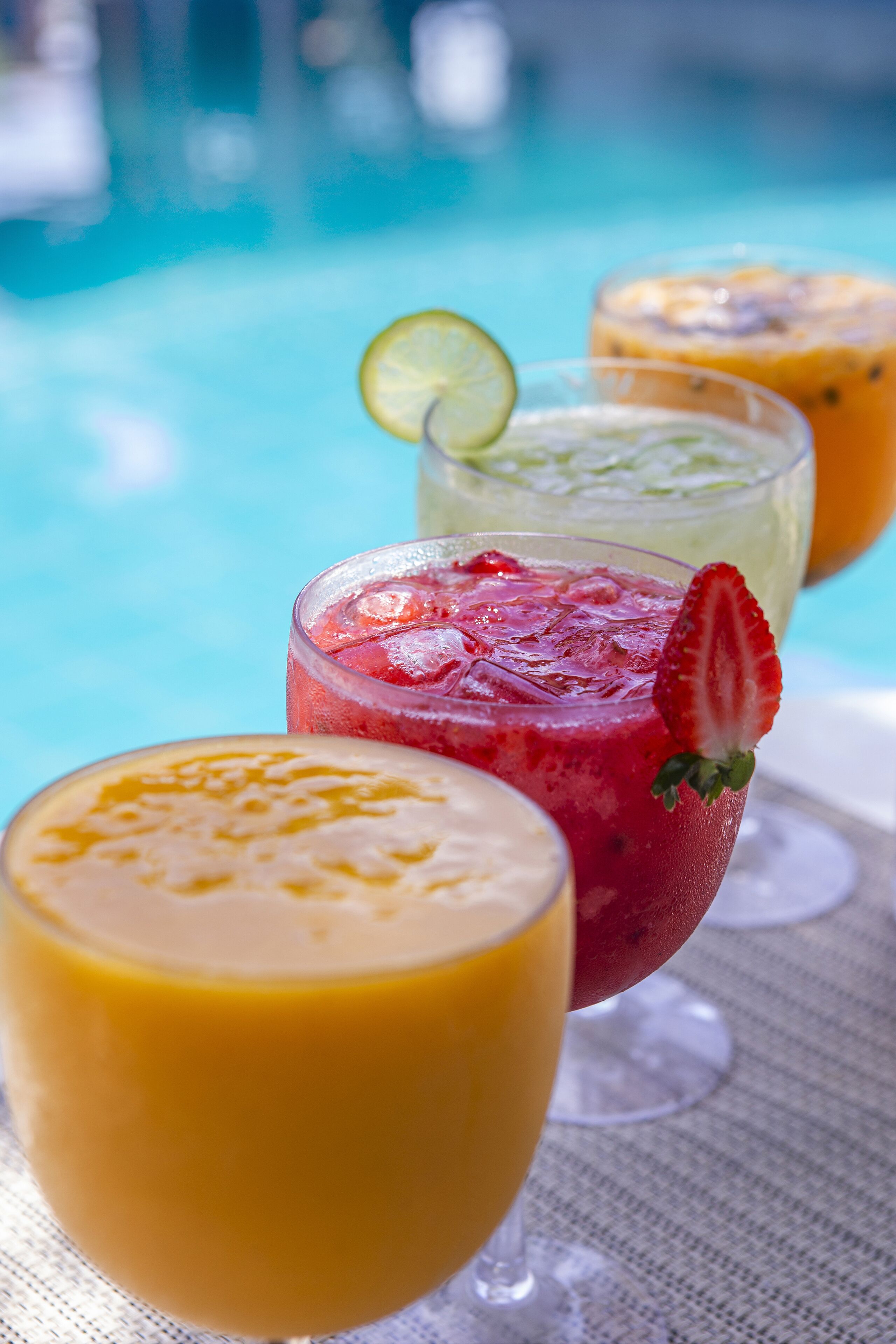 Tres vasos de bebidas con hielo y adornos, sobre una mesa cerca de una piscina.