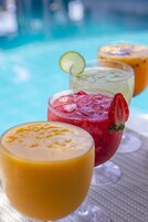 Tres vasos de bebidas con hielo y adornos, sobre una mesa cerca de una piscina.