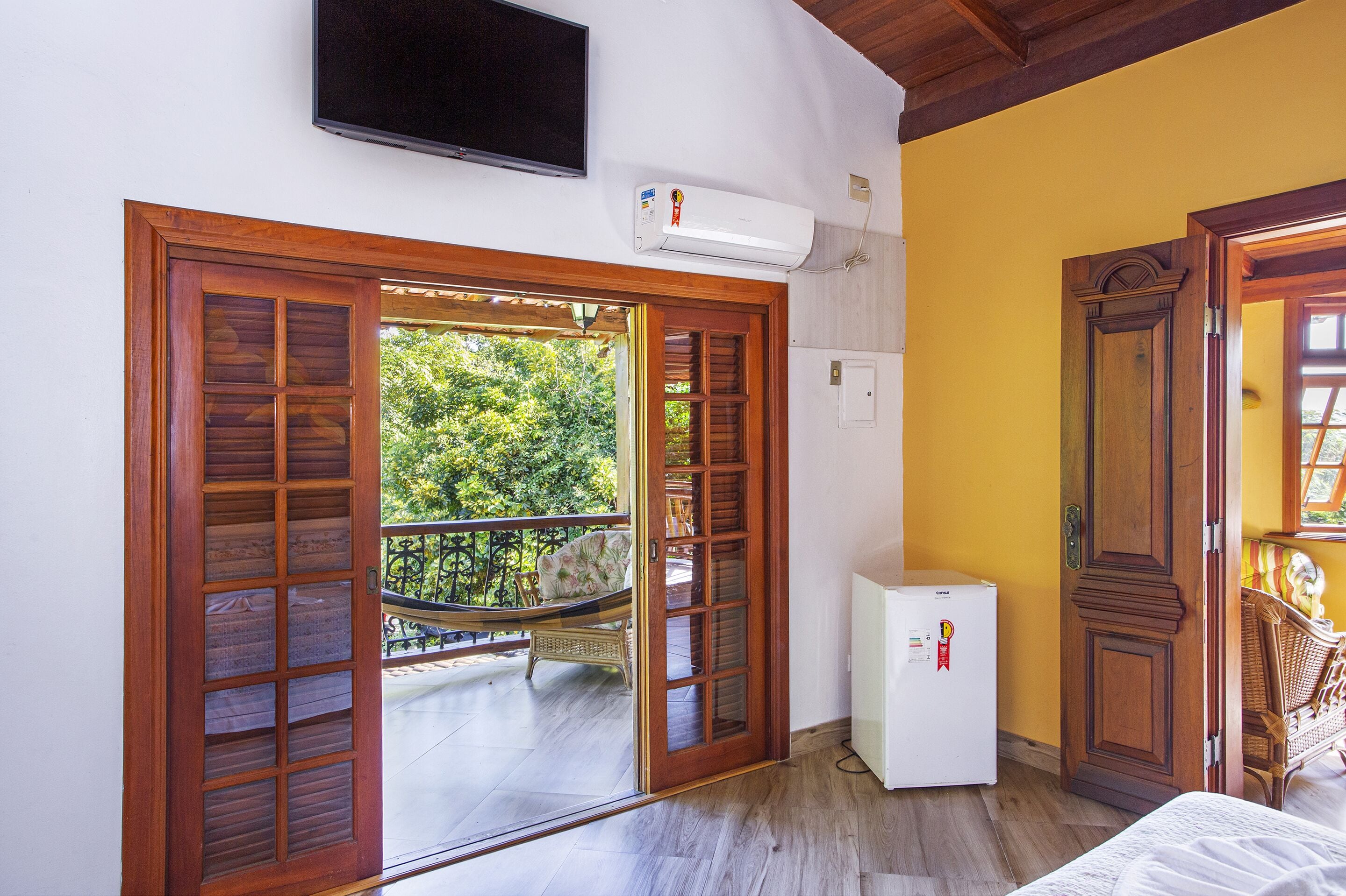 Um quarto com porta de madeira, um pequeno frigorífico branco e uma televisão montada na parede.