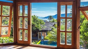 Habitación con amplios ventanales que dan a una piscina, plantas tropicales y una construcción con techo de paja.