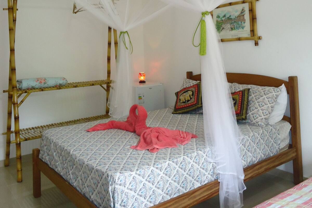 A room with a bed under a mosquito net, a bedside table with a lamp, and a wall-mounted picture.