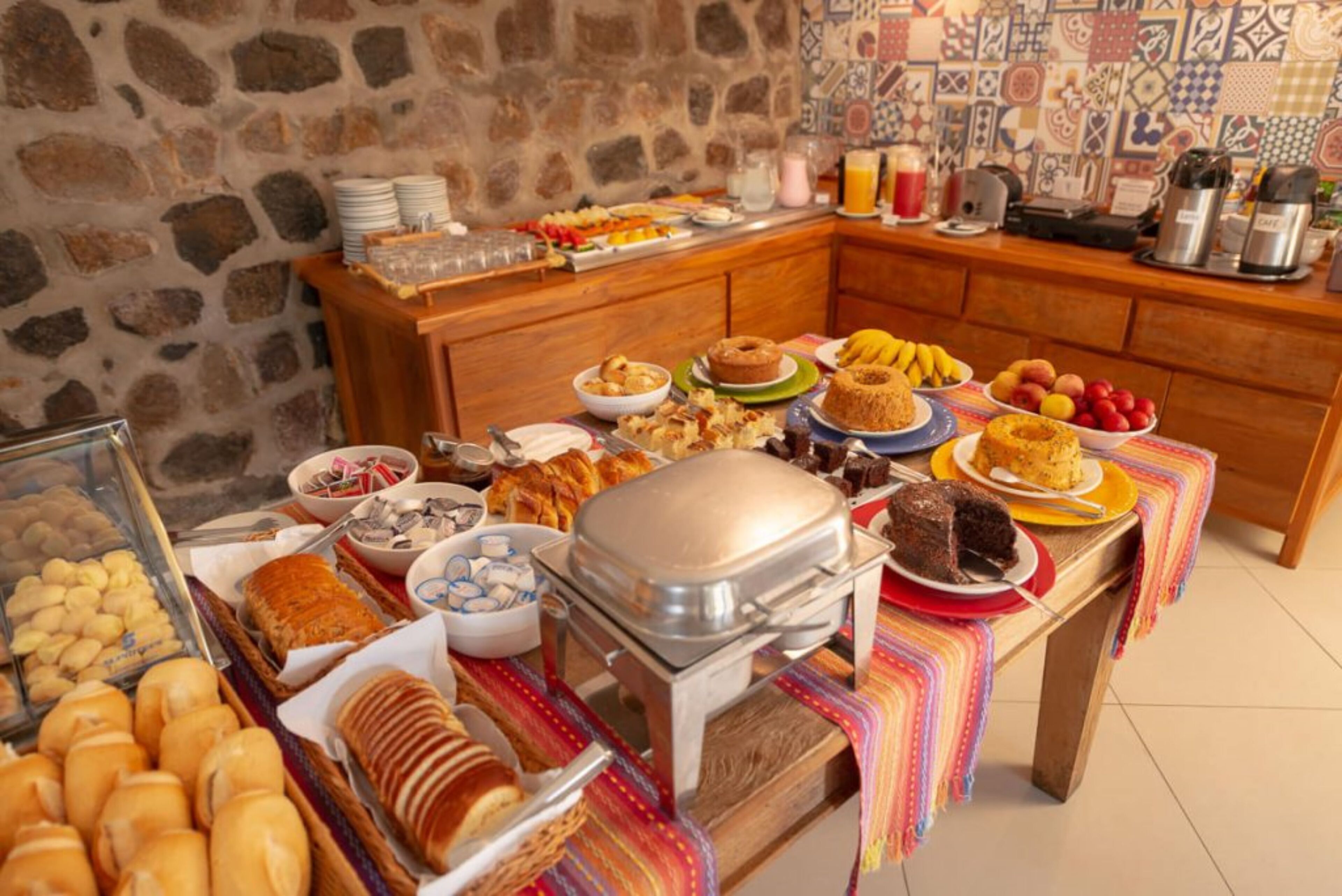 Un bufé con diferentes opciones de comida, entre ellas pasteles, frutas y un pastel, dispuesto sobre una mesa de madera con un mantel decorativo de colores.