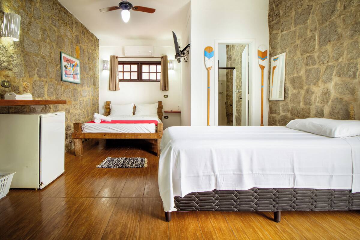 A bedroom with a wooden bed, a small desk, a refrigerator, and a stone wall.