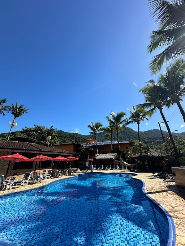 A swimming pool with lounge chairs and umbrellas, surrounded by palm trees and a mountainous backdrop. - Hotel Pousada dos Condes (São Sebastião)