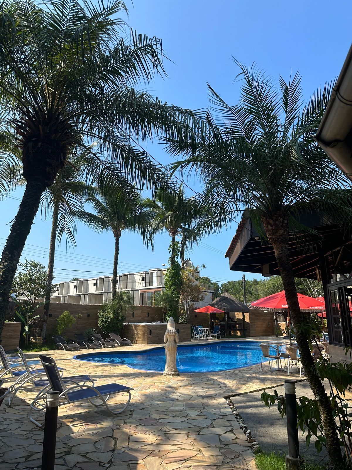 Zona de piscina con sillas de descanso, terraza de baldosas de piedra, palmeras y un edificio al fondo.