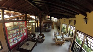Amplia habitación con techo alto, vigas de madera y piso de baldosas. Hay varias sillas y mesas de madera, y un ventanal con vistas a la vegetación exterior.