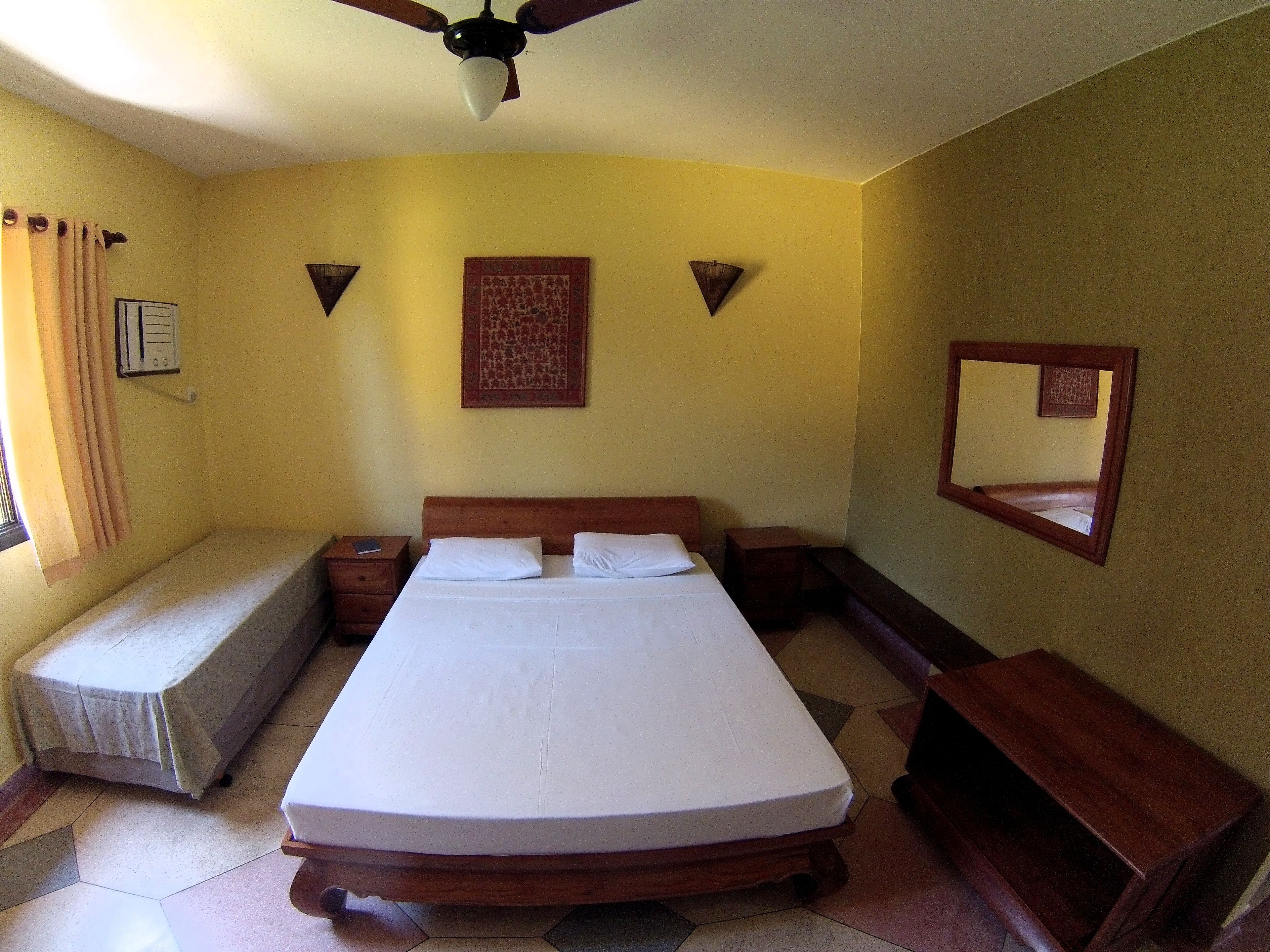 Habitación de hotel con una cama individual, una mesita de noche, un ventilador de techo, una ventana con cortinas y un espejo colgado en la pared.