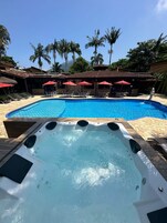 A swimming pool with lounge chairs and umbrellas, a hot tub, and palm trees in the background.