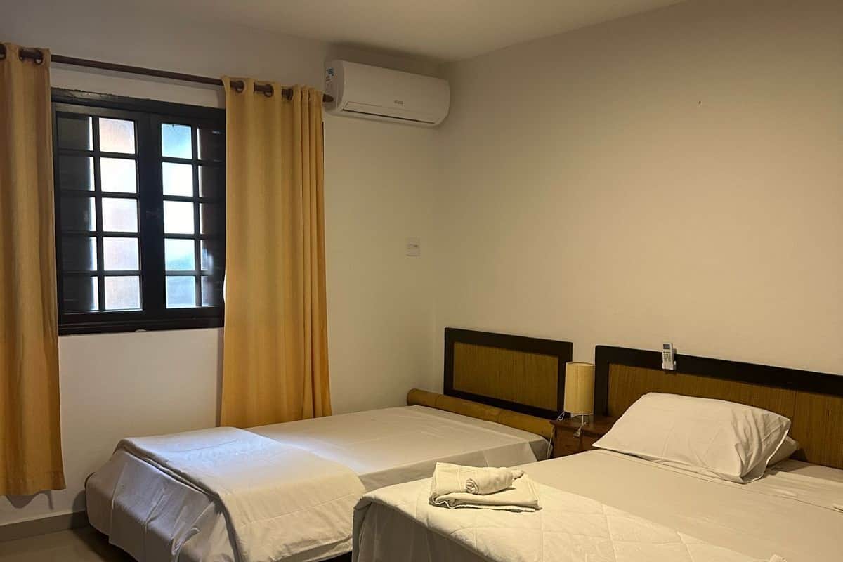 A hotel room with two single beds, a window with curtains, an air conditioning unit, and a bedside lamp.
