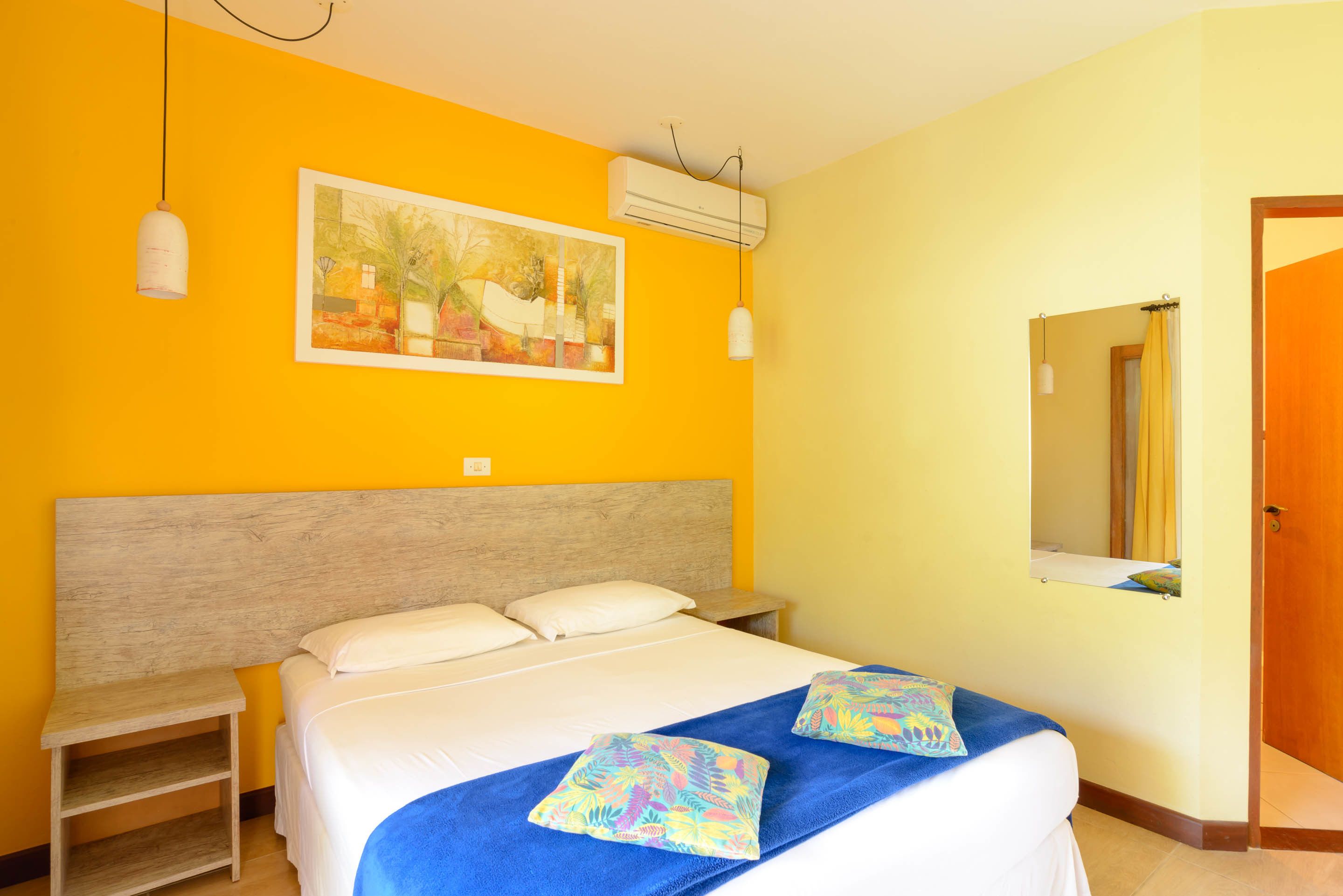 A hotel room with a bed, a nightstand, a painting on the wall, and an air conditioning unit.