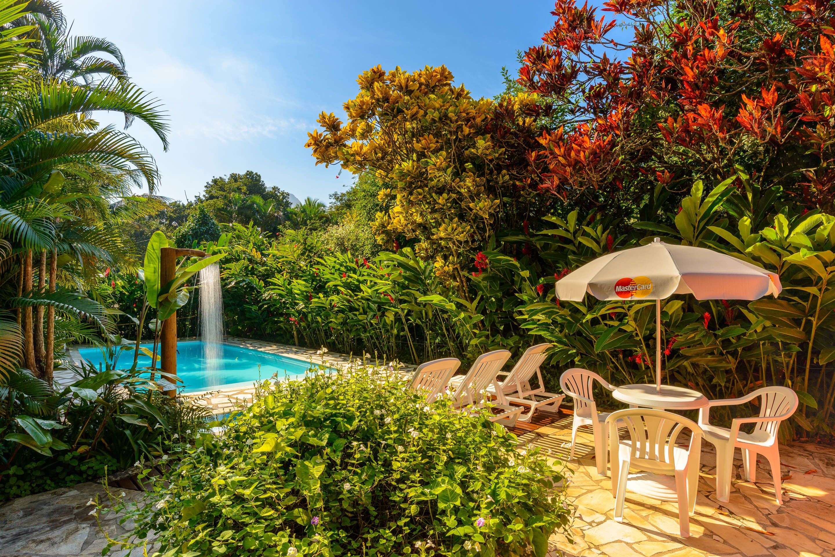 A pool area with a waterfall, surrounded by lush greenery and white plastic chairs and tables.