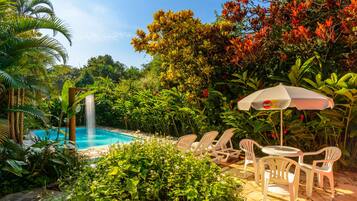 A pool area with a waterfall, surrounded by lush greenery and white plastic chairs and tables.