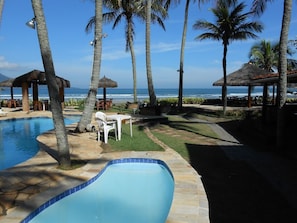 A pool area with a curved edge, surrounded by palm trees and a view of the ocean. - Barequeçaba Praia Hotel (São Sebastião)