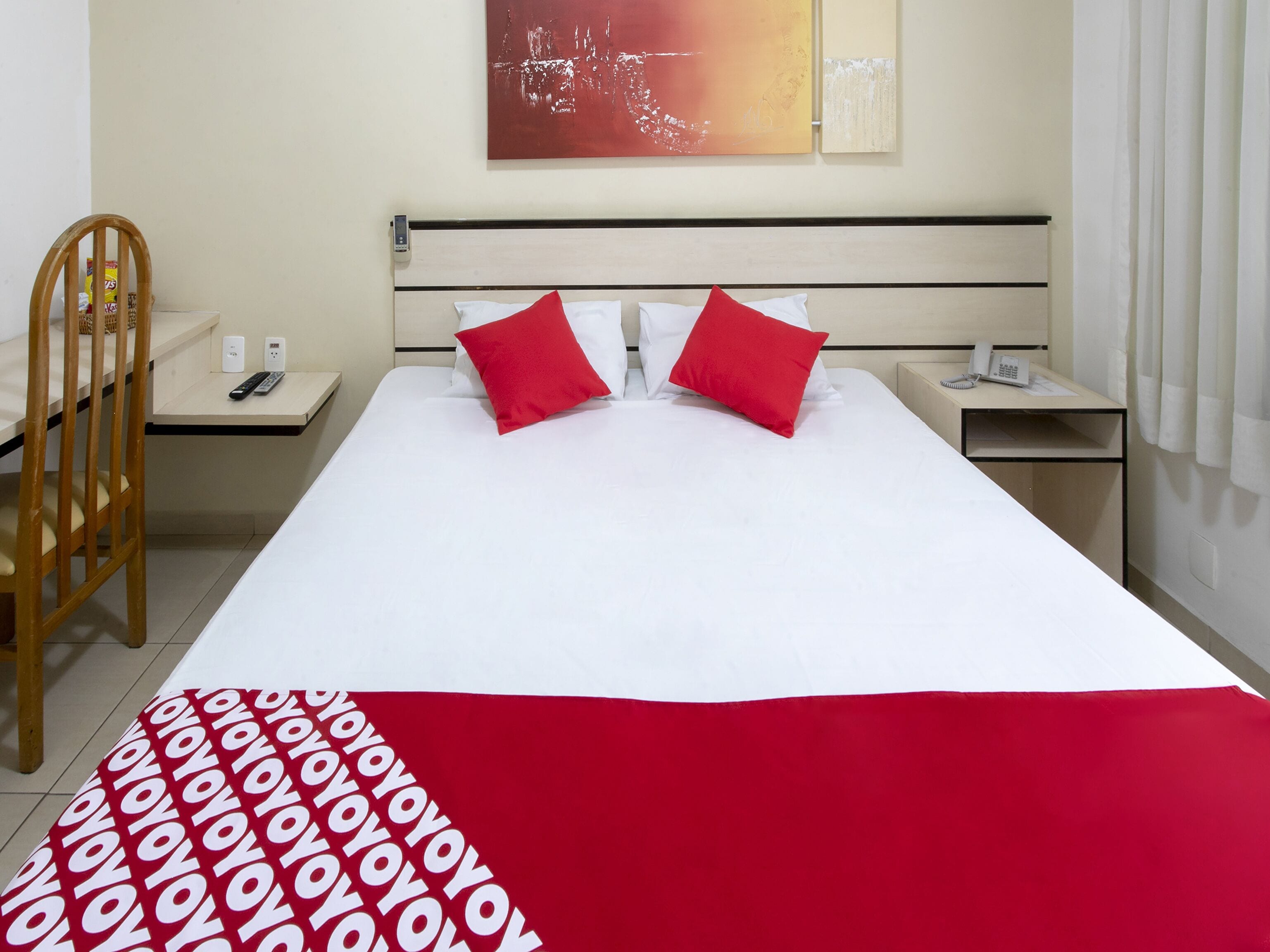 A hotel room with a large bed, two red pillows, a white bedspread, a red and white patterned blanket, a wooden chair, a bedside table with a phone, and a wall art piece.