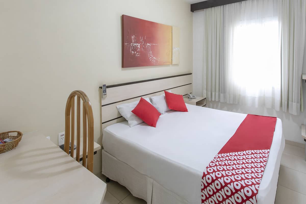 Habitació d'hotel amb un llit, una taula de nit, una cadira, un telèfon fixat a la paret i una finestra amb cortines.