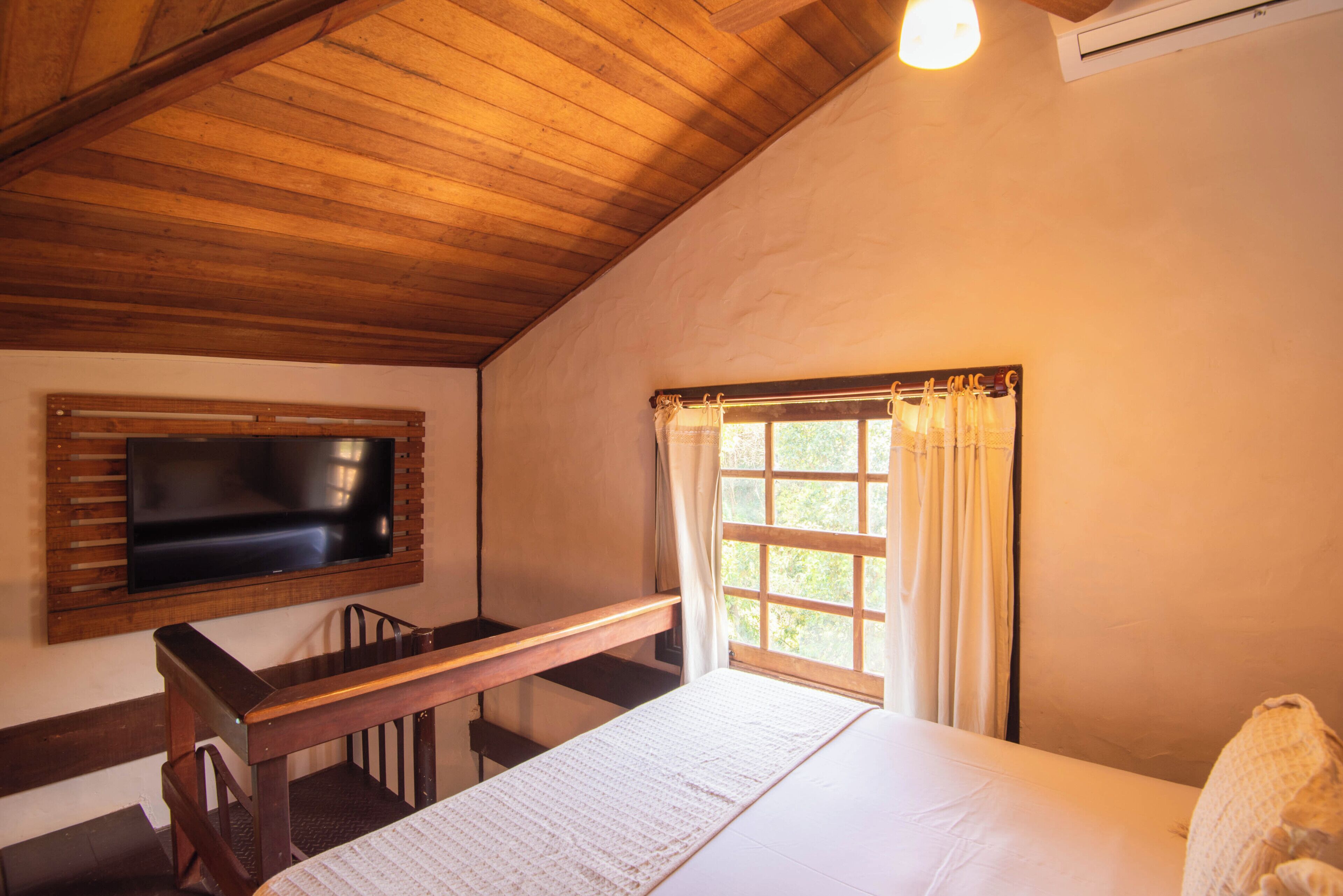 A room with a wooden ceiling, a bed with a white mattress, a wooden desk with a chair, a television mounted on the wall, and a window with curtains.